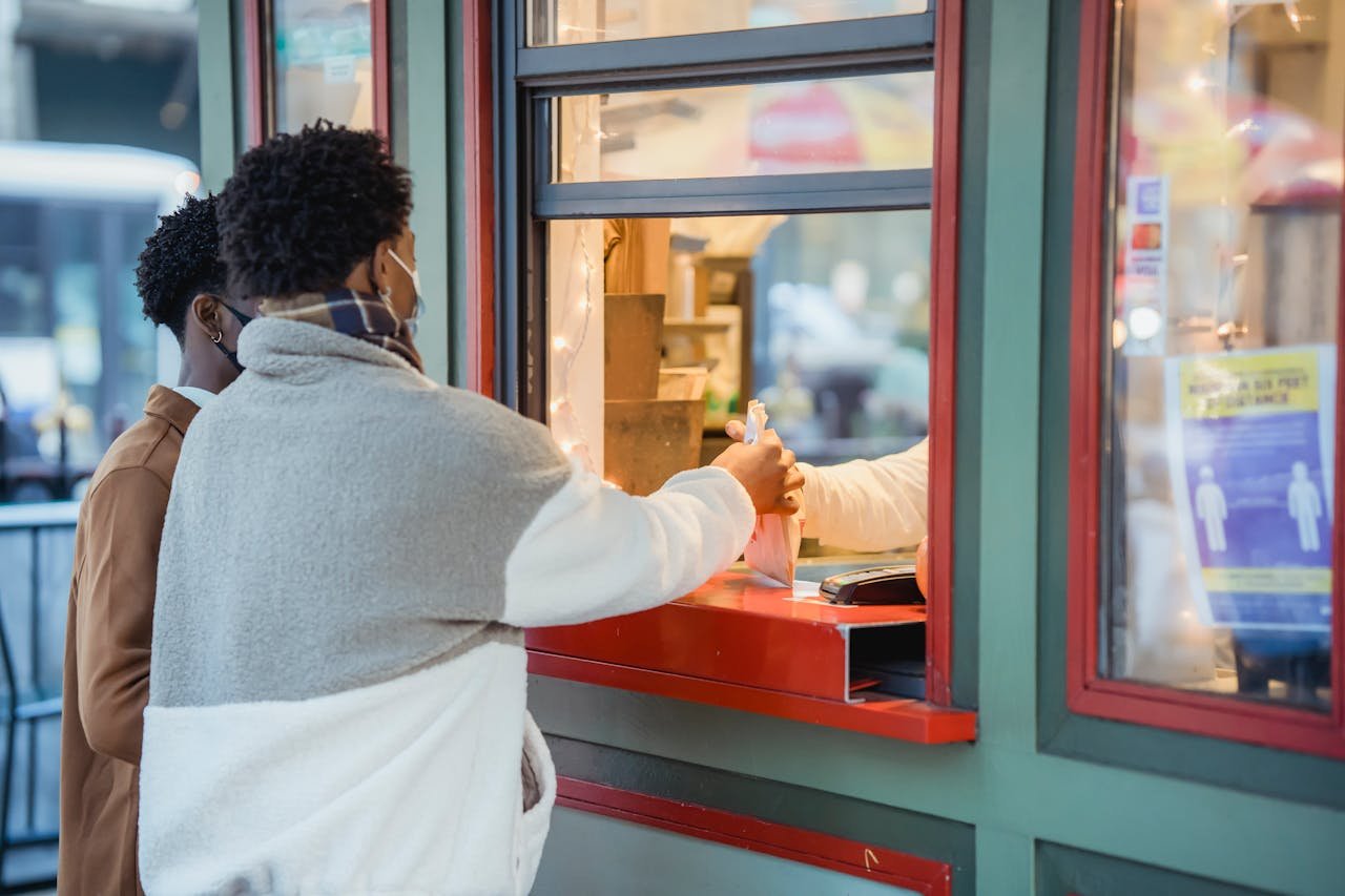 Side view of anonymous African American male couple in medical masks buying food on street while standing near window of store